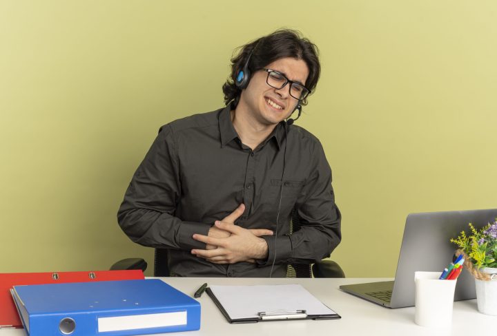 young annoyed office worker man on headphones in optical glasses sits at desk with office tools using laptop puts hands on belly isolated on green background with copy space